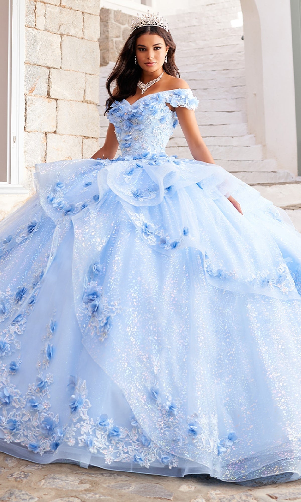A woman wearing the Quinceanera Dress PR30113 By Princesa—sparkling light blue, off-the-shoulder, with floral details and a tiara—stands on a stone staircase outdoors.